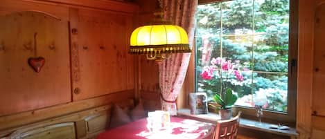 A wooden room with a dining table, a hanging lamp, a window with a view of flowers, and a heart-shaped decoration on the wall.