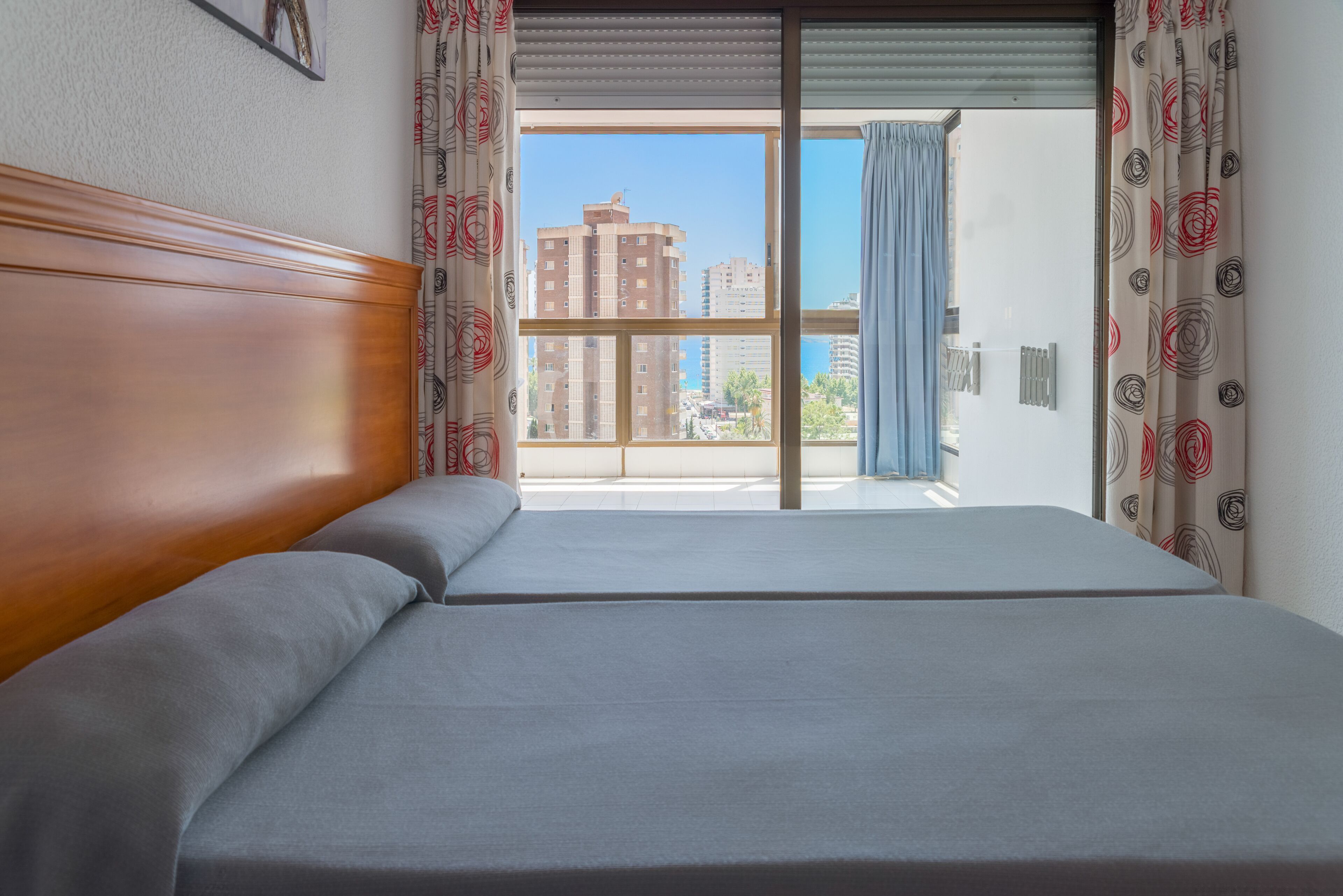 A hotel room with a large bed, wooden headboard, and a view of tall buildings through a window with patterned curtains.