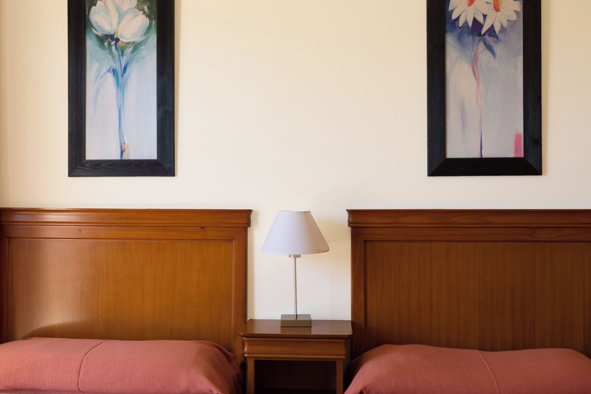 A hotel room with two beds, a nightstand with a lamp, and two framed floral paintings on the wall.