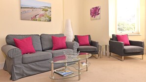A living room with a grey sofa, two armchairs, a glass coffee table, and a painting of a beach scene. - Cavendish Apartment (Eastbourne)