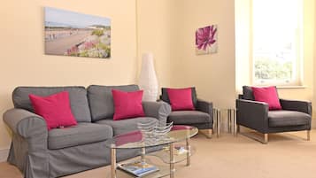 Uma sala de estar com um sofá cinzento, duas poltronas, uma mesa de centro de vidro e um quadro de uma praia.