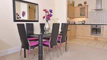 Uma sala de jantar com mesa de vidro e cadeiras pretas, um vaso com flores e uma cozinha ao fundo.