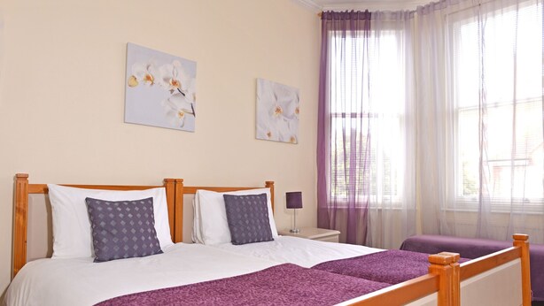A double bed with a wooden frame, two pillows, a purple blanket, and a nightstand with a lamp. - Cavendish Apartment (Eastbourne)