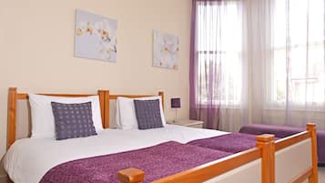 Uma cama de casal com estrutura de madeira, duas almofadas, um cobertor roxo e uma mesa de cabeceira com abajur.