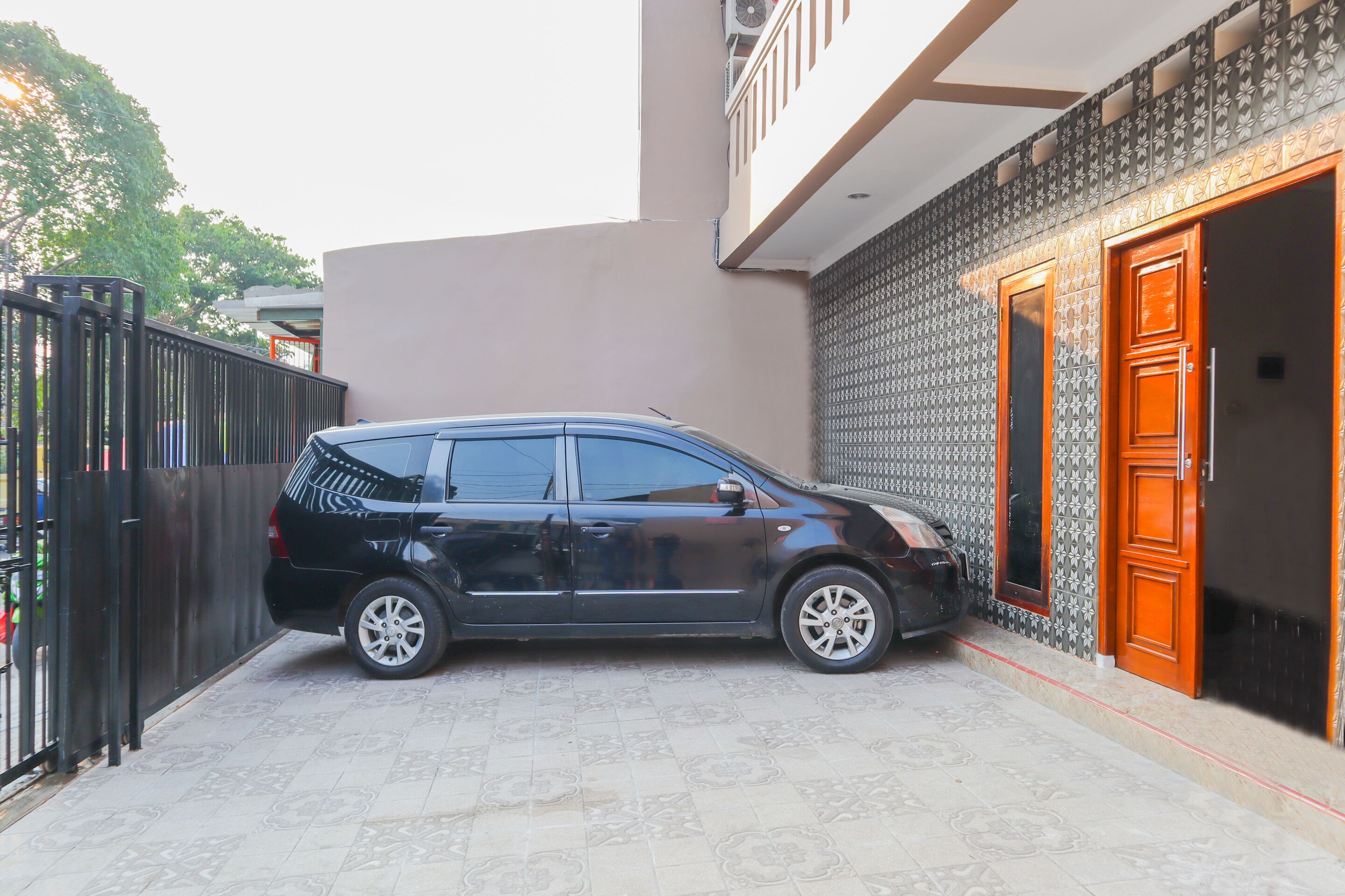 A black car parked in a driveway with a patterned tiled floor, a black gate, and a building with patterned walls and a wooden door.
