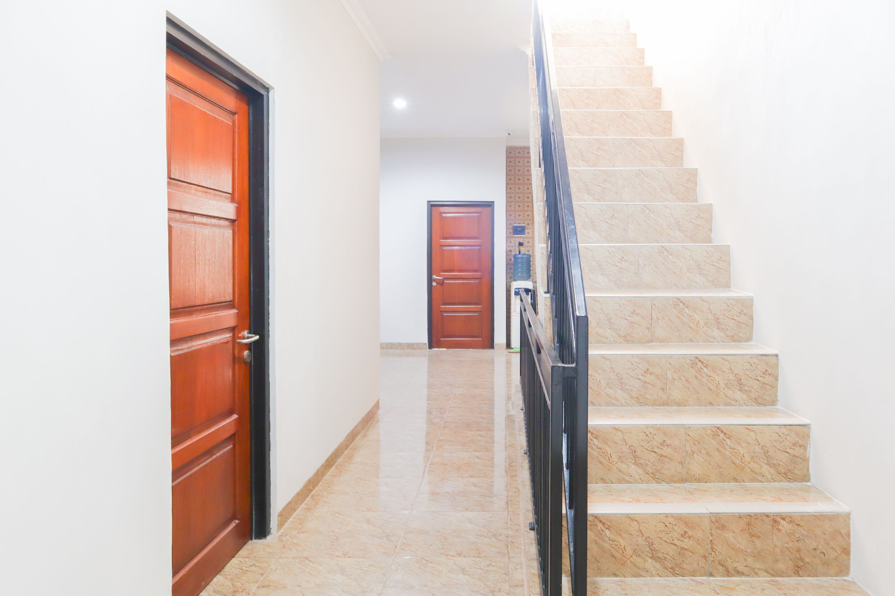 Um corredor com uma porta de madeira à esquerda, uma escadaria de mármore à direita e outra porta de madeira no final.
