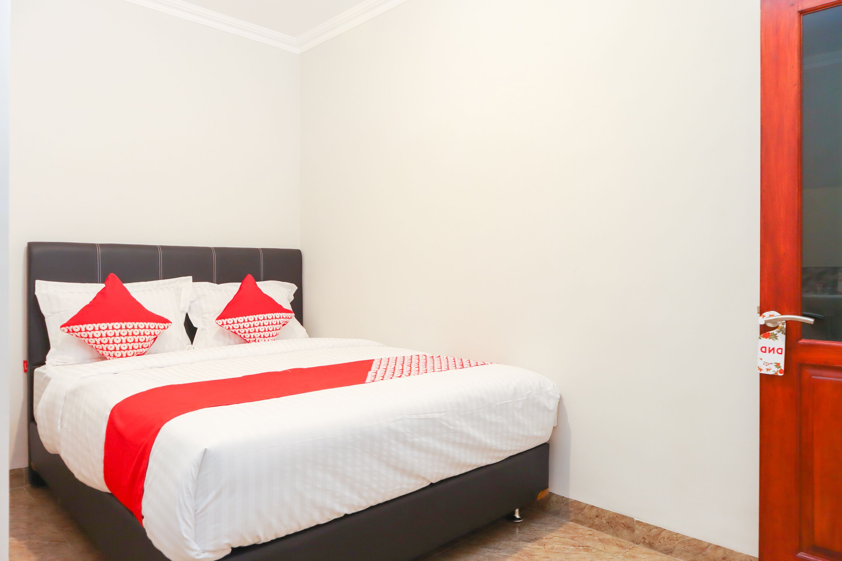 A neatly made bed with white and red bedding, a dark headboard, and a wooden door with a sign.