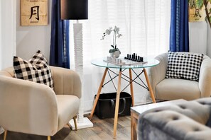 A living room with two beige armchairs, a glass coffee table, a chessboard, and a plant.