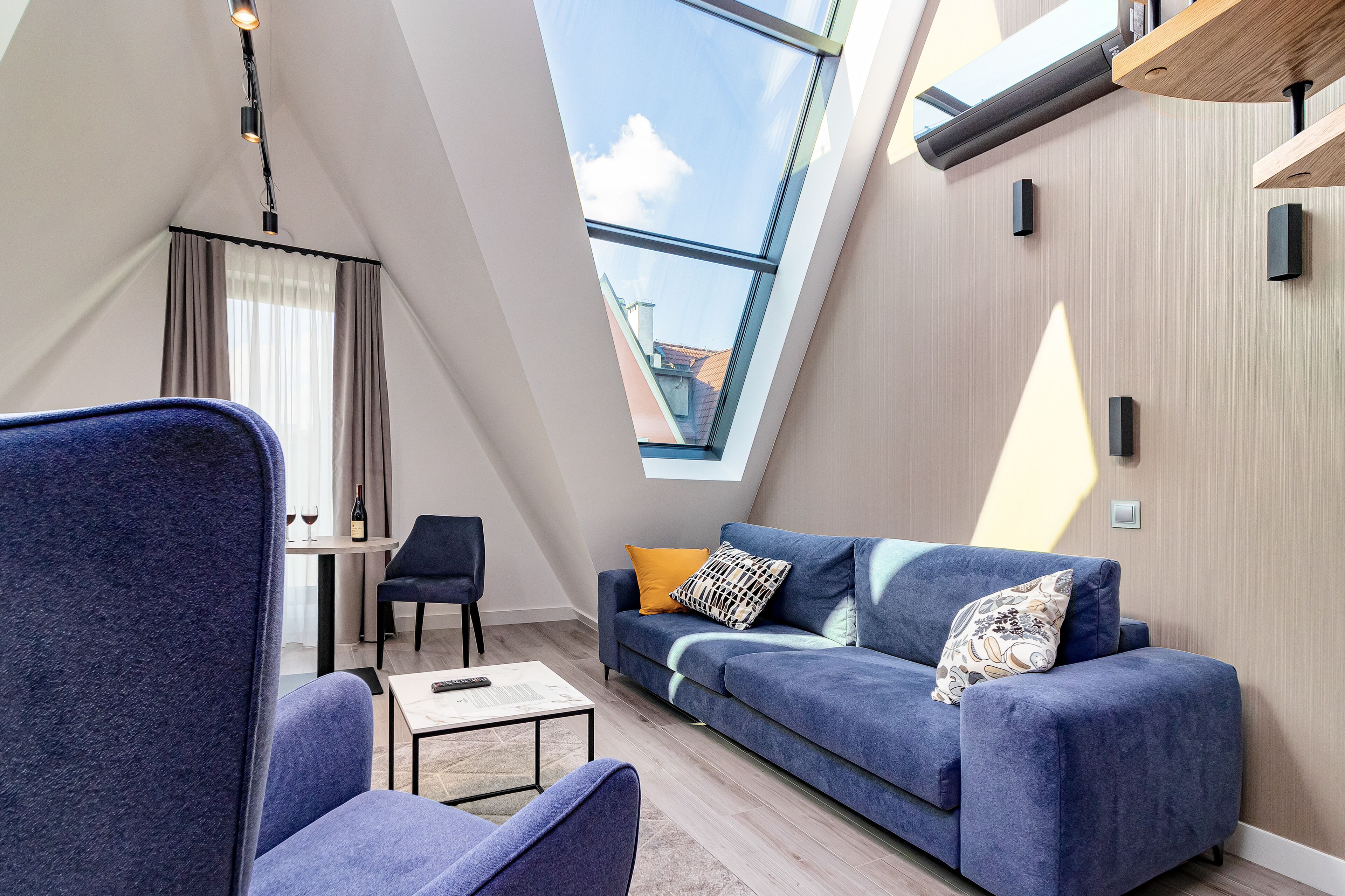 A modern living room with a blue sofa, a coffee table, and a dining area with a table and chairs. A skylight provides natural light.