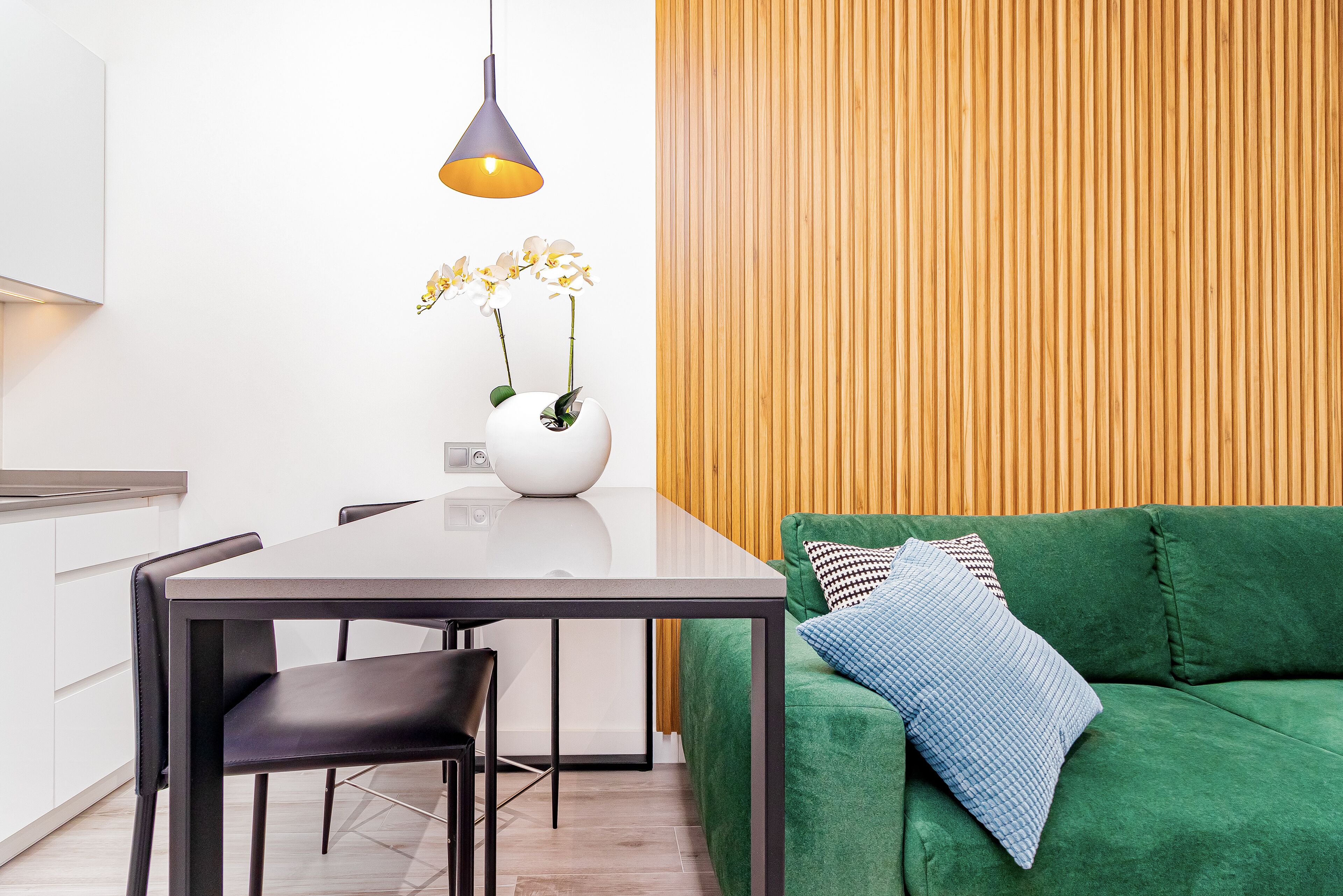 A modern living space with a dining table, chairs, a green sofa with patterned cushions, a white vase with flowers, and a pendant light.