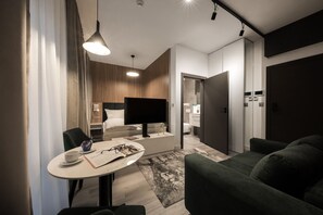 Quarto de hotel moderno com cama, televisão, área de refeições com mesa e cadeiras e um sofá verde.