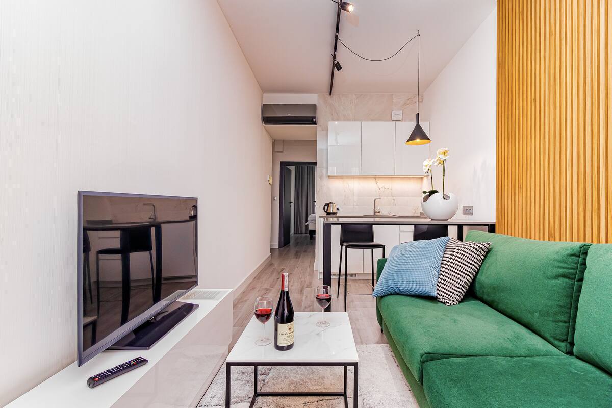 Een moderne woonkamer met een flatscreen televisie, een groene bank, een bijzettafeltje met wijnglazen en een fles, en een keuken op de achtergrond.