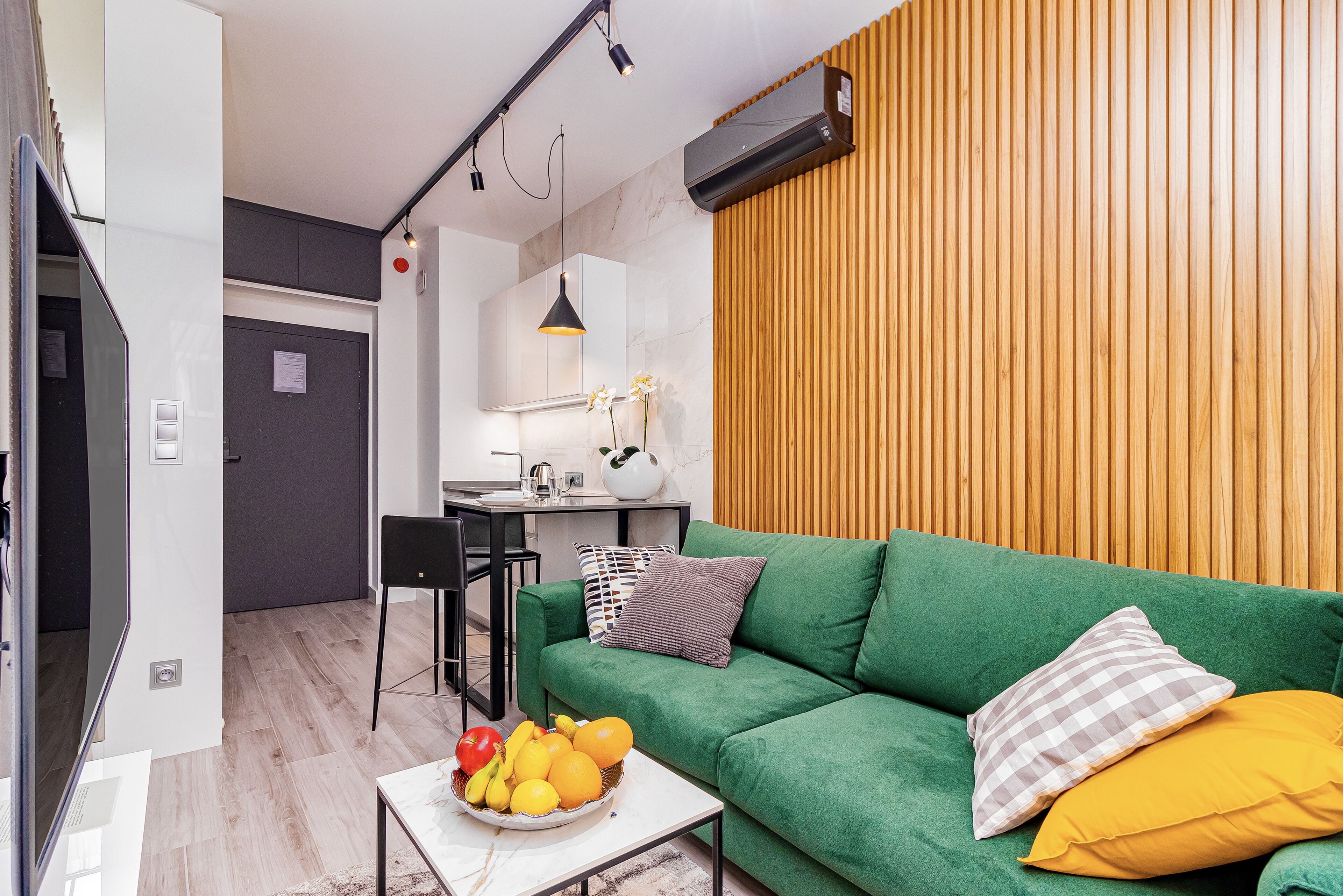 Ein modernes Wohnzimmer mit einem grünen Sofa, einem Couchtisch mit Früchten, einer Holzvertäfelung und einer Küche im Hintergrund.