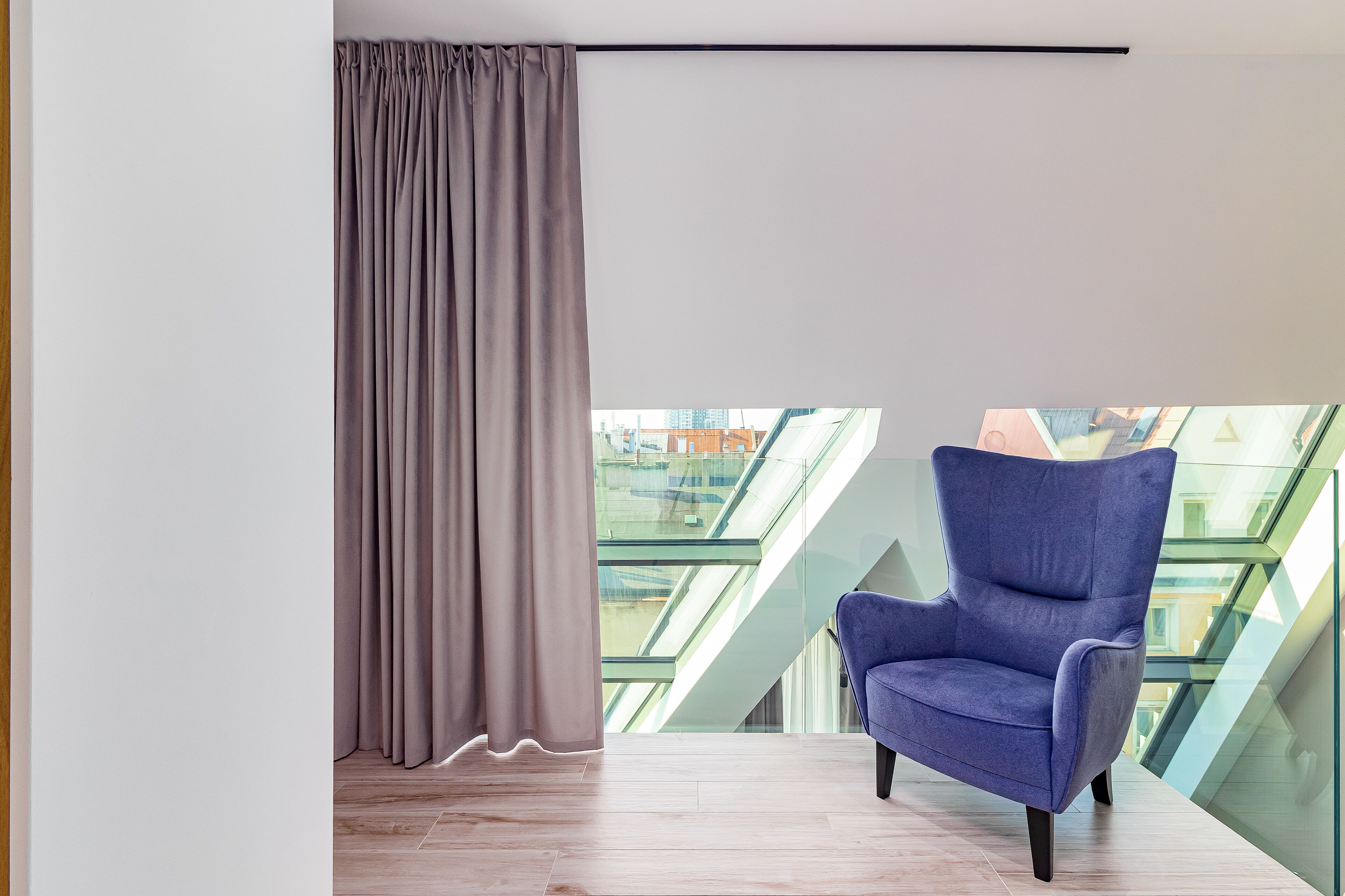Una habitación moderna con techo alto, un ventanal con vistas a un paisaje urbano, un sillón azul y una cortina.