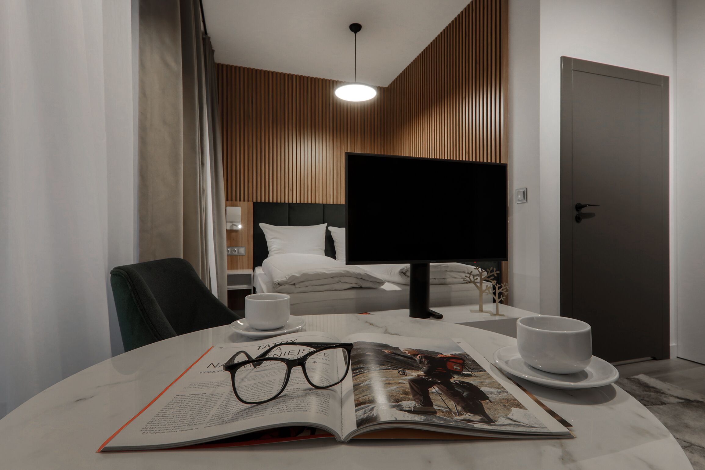 Habitación de hotel con cama, televisor, escritorio con una revista, vasos y dos tazas.