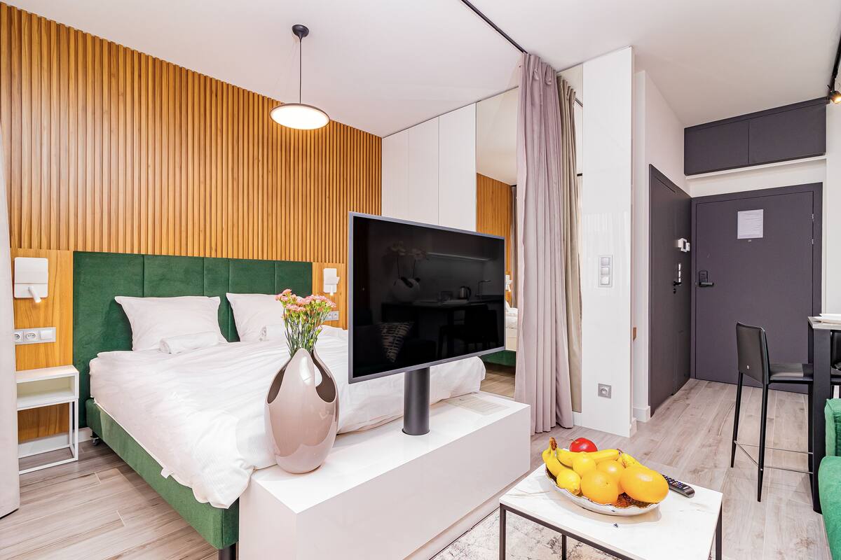 Een moderne hotelkamer met een groen bekleden hoofdeinde, een flatscreen televisie, een vaas met bloemen en een schaal fruit op het nachtkastje.