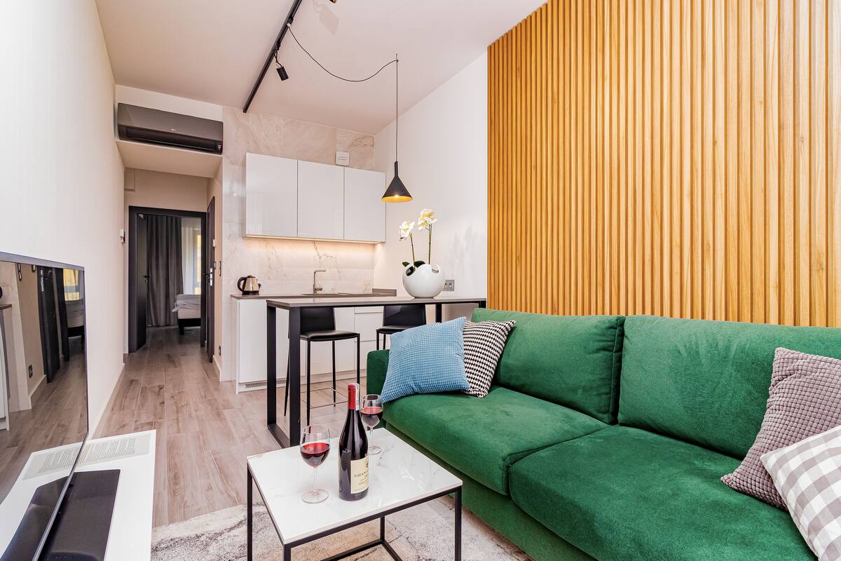 Een moderne woonkamer met een groene bank, een klein tafeltje met een fles wijn, en een keuken op de achtergrond.