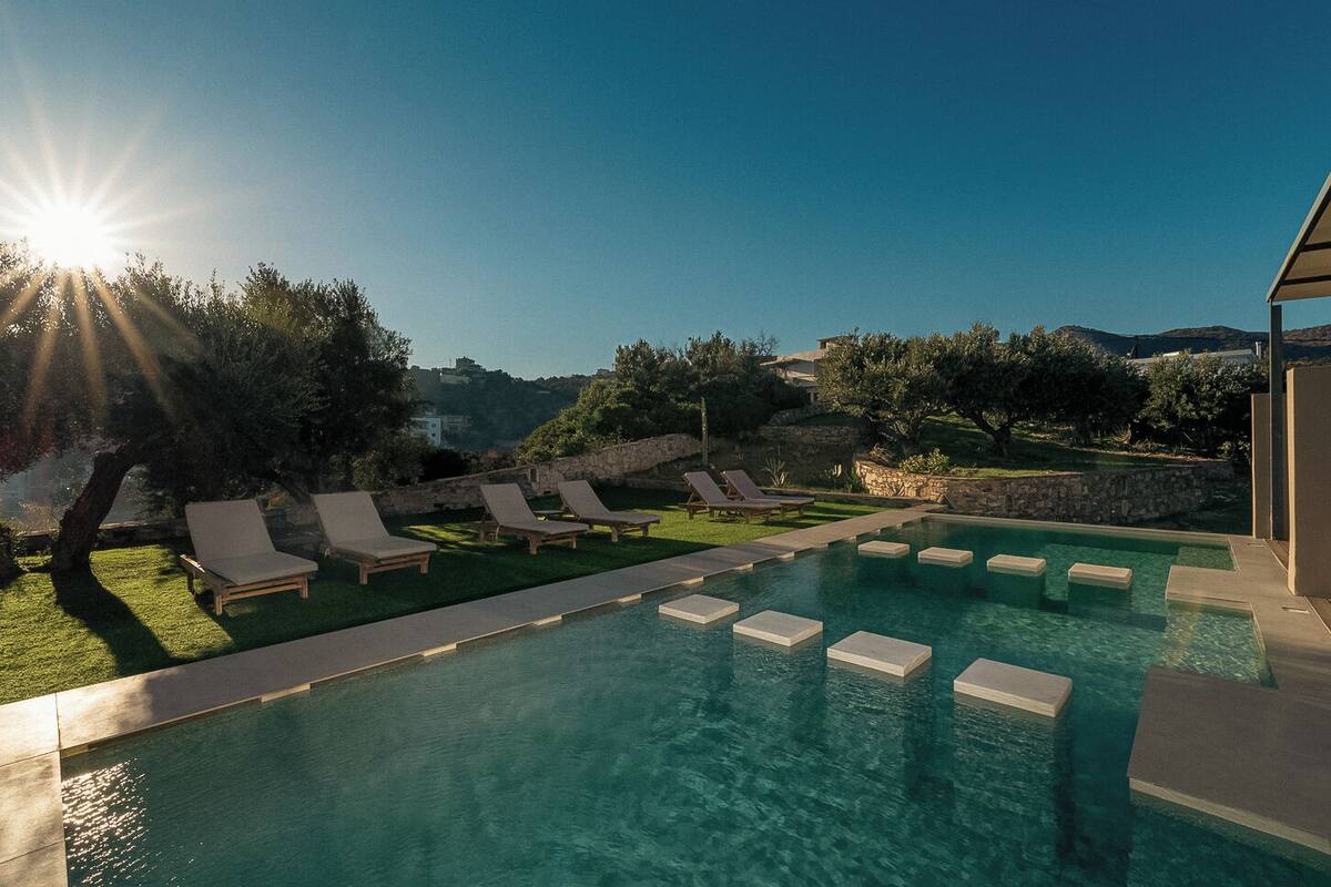 Un área de piscina con agua iluminada por el sol, sillones de descanso y un muro de piedra.
