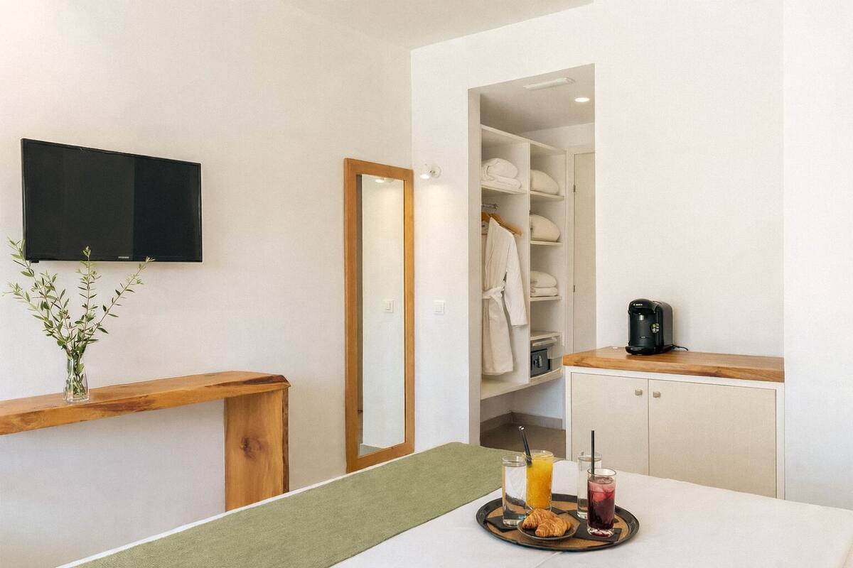 Habitación de hotel con un banco de madera, televisor de pantalla plana, espejo, armario con batas y bandeja de desayuno con croissants y bebidas.