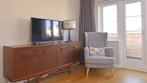 A living room with a wooden cabinet, a flat-screen TV, a lamp, and a grey armchair with floral cushions. - Dress Circle (Eastbourne)
