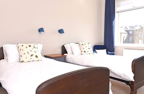 A hotel room with two single beds, a wooden headboard, a nightstand, a blue chair, and a window with a view of buildings. - Dress Circle (Eastbourne)
