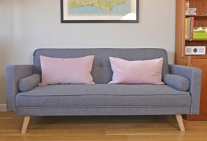 A grey sofa with two pink cushions, a framed map on the wall, and a bookshelf with books and a radio. - Dress Circle (Eastbourne)