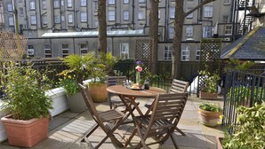 Eine Terrasse mit einem runden Tisch und vier Holzstühlen, umgeben von Topfpflanzen und einem Metallzaun.