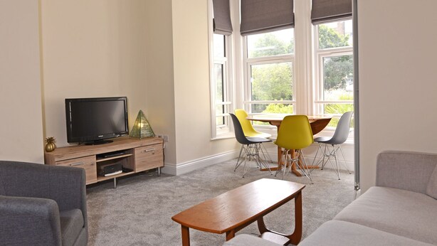 A modern living room with a flat-screen TV, a wooden entertainment unit, a dining table with chairs, and a sofa.