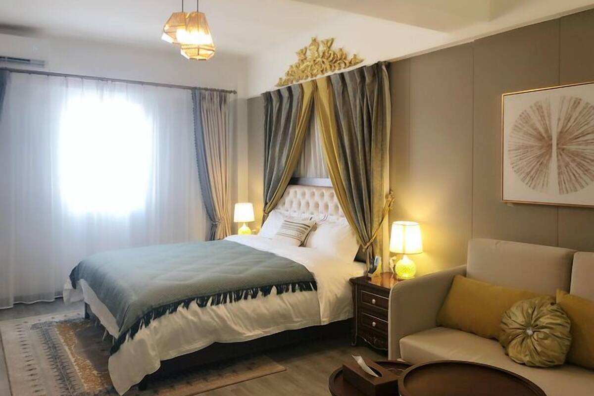 A hotel room with a large bed, a sofa, a nightstand with lamps, and a wall-mounted artwork.