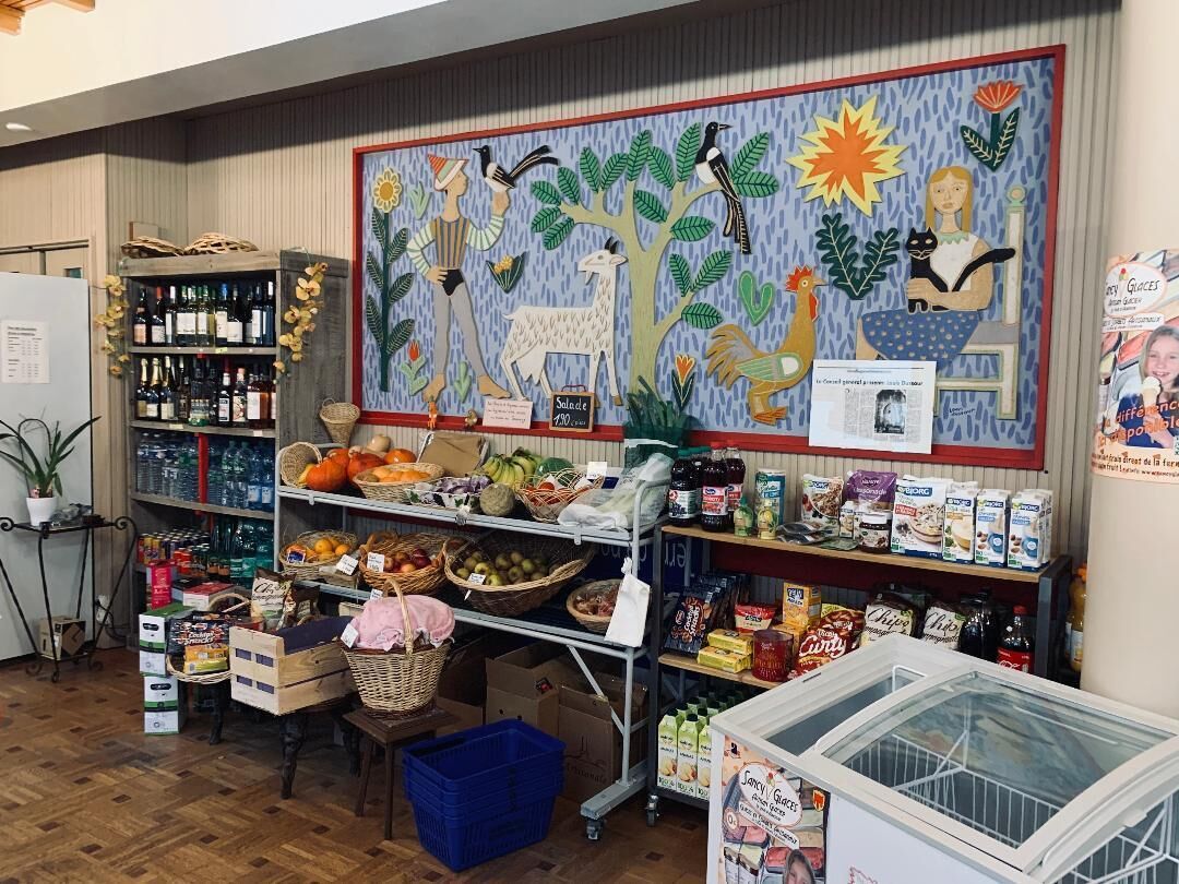 Interior de un supermercado con estantes llenos de diversos productos, una exhibición de frutas y verduras frescas, y un mural colorido en la pared.