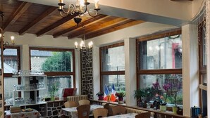 Ein Restaurant mit Holzbalken, Steinwänden und großen Fenstern. Die Tische sind mit Stühlen gedeckt, und auf dem Fensterbrett stehen Blumentöpfe.