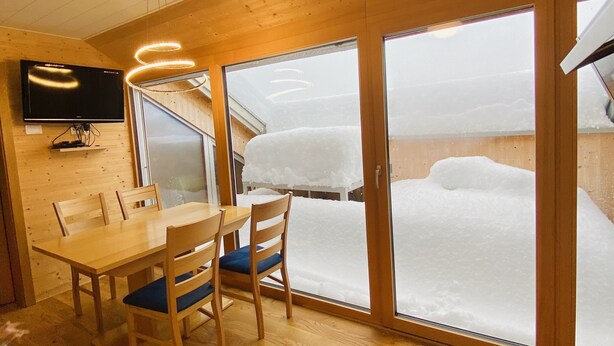 A wooden interior with a dining table and chairs, a television mounted on the wall, and large glass doors leading to a snowy outdoor area. - Rose house in Schwarzenberg (Schwarzenberg)