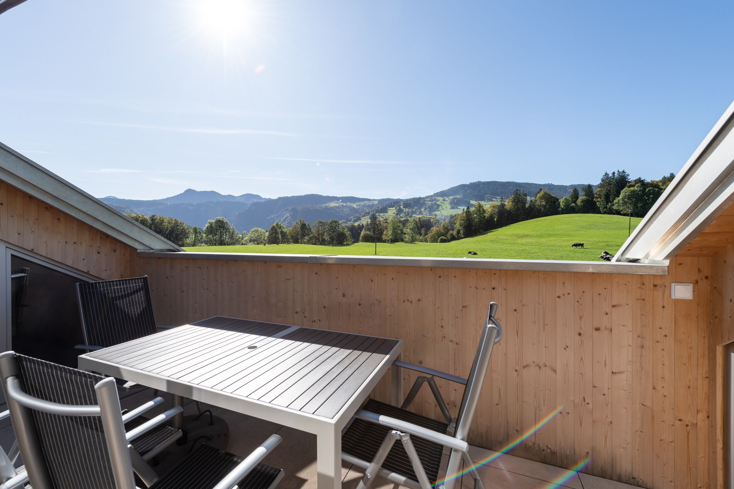 A wooden deck with a table and chairs, overlooking a green field and mountains.
