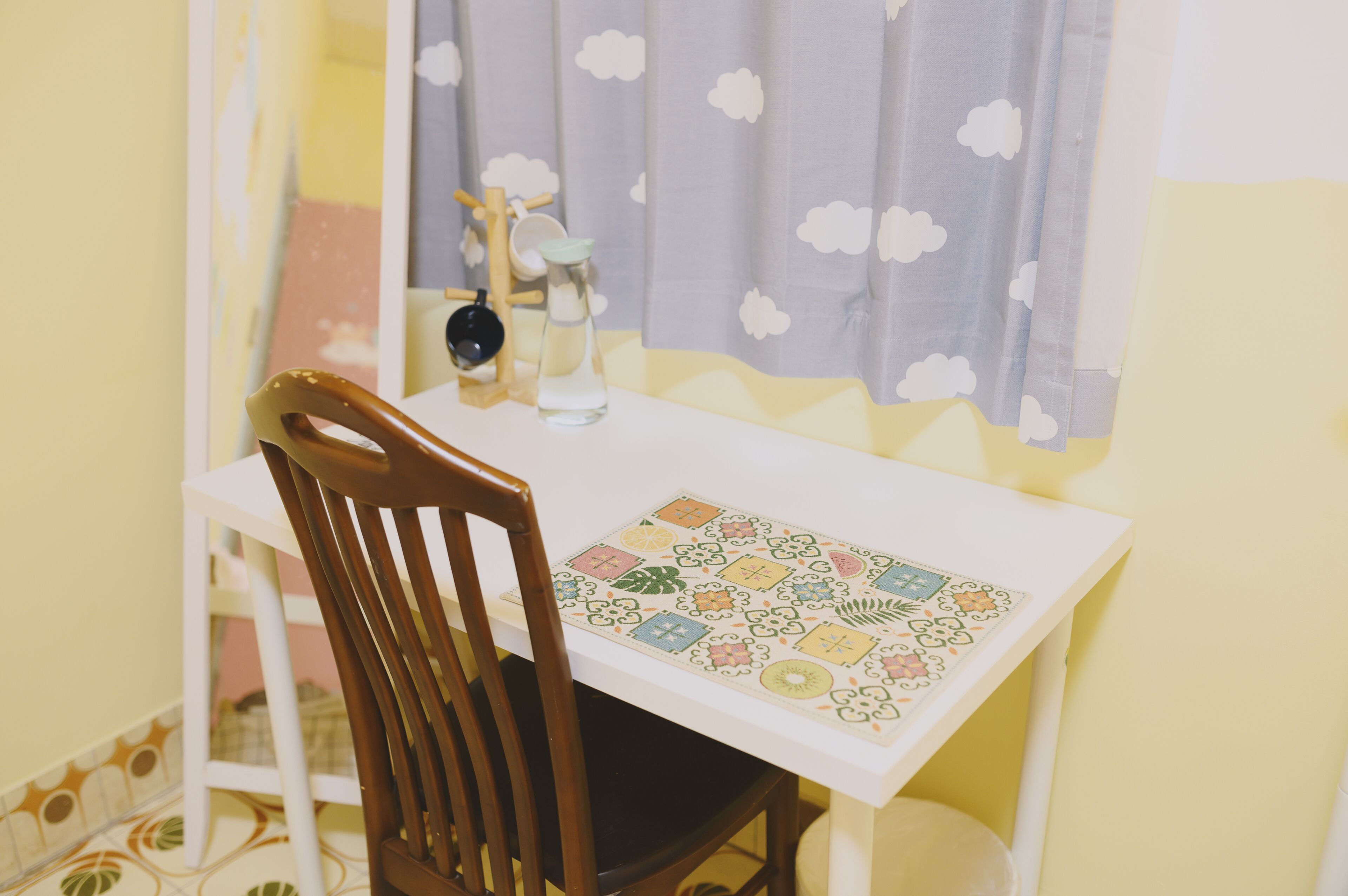 A small dining table with a patterned tablecloth, a wooden chair, a vase with a black object, and a curtain with a cloud pattern.