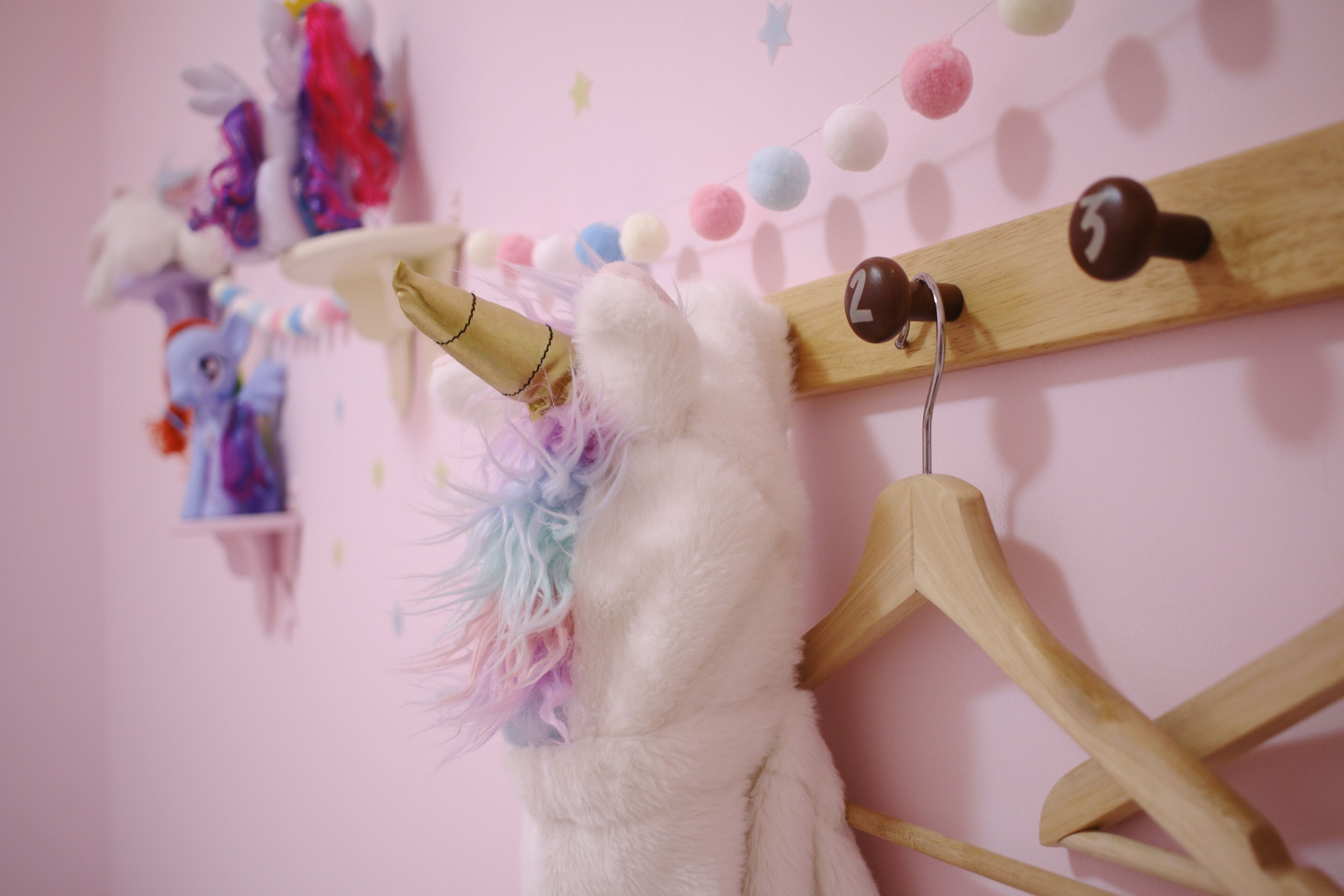 A unicorn plush toy on a wooden hanger against a pink wall with a string of colorful pom-poms and a small shelf with plush toys.