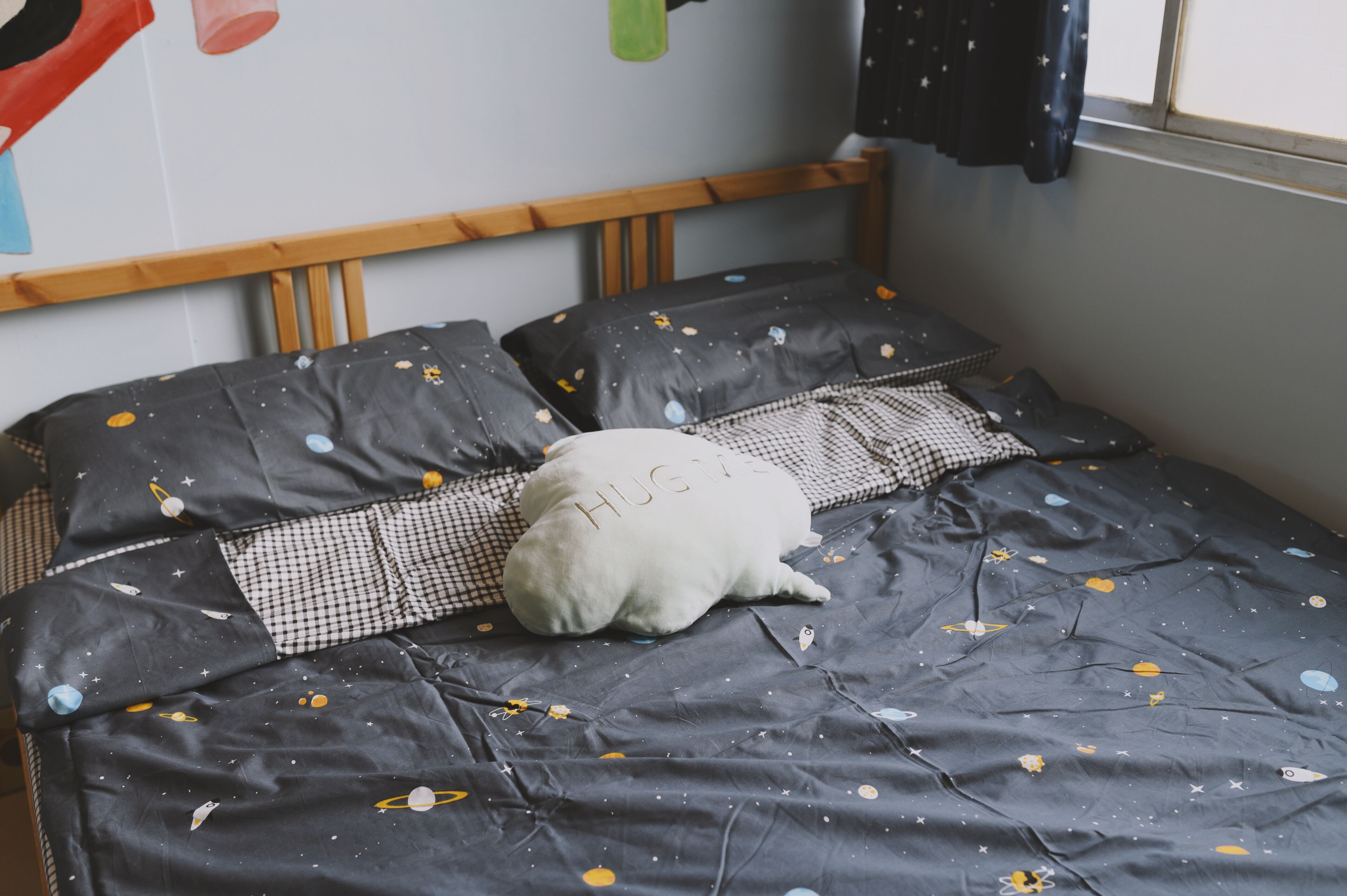 A bed with a space-themed bedspread, a pillow with the word "HUG" on it, and a checkered pillow.