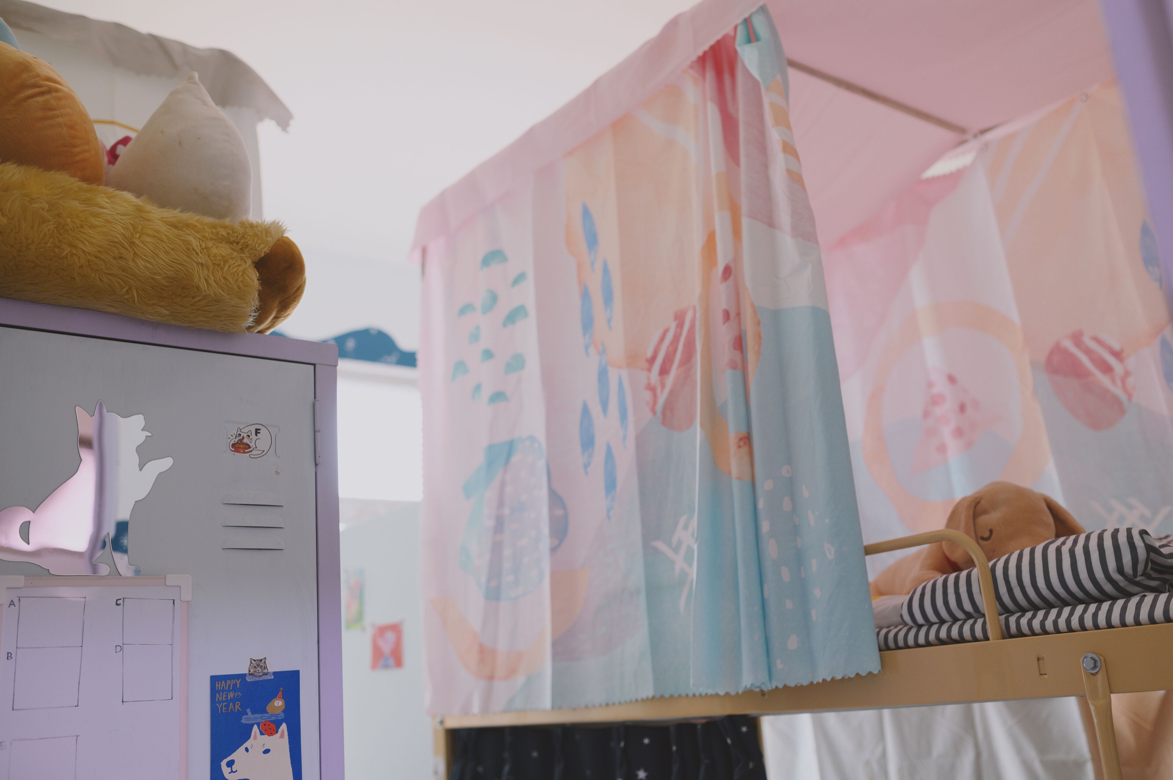 A child's bunk bed with a striped blanket and a stuffed animal, a whiteboard with drawings and a cat sticker, and a pink curtain with abstract patterns.