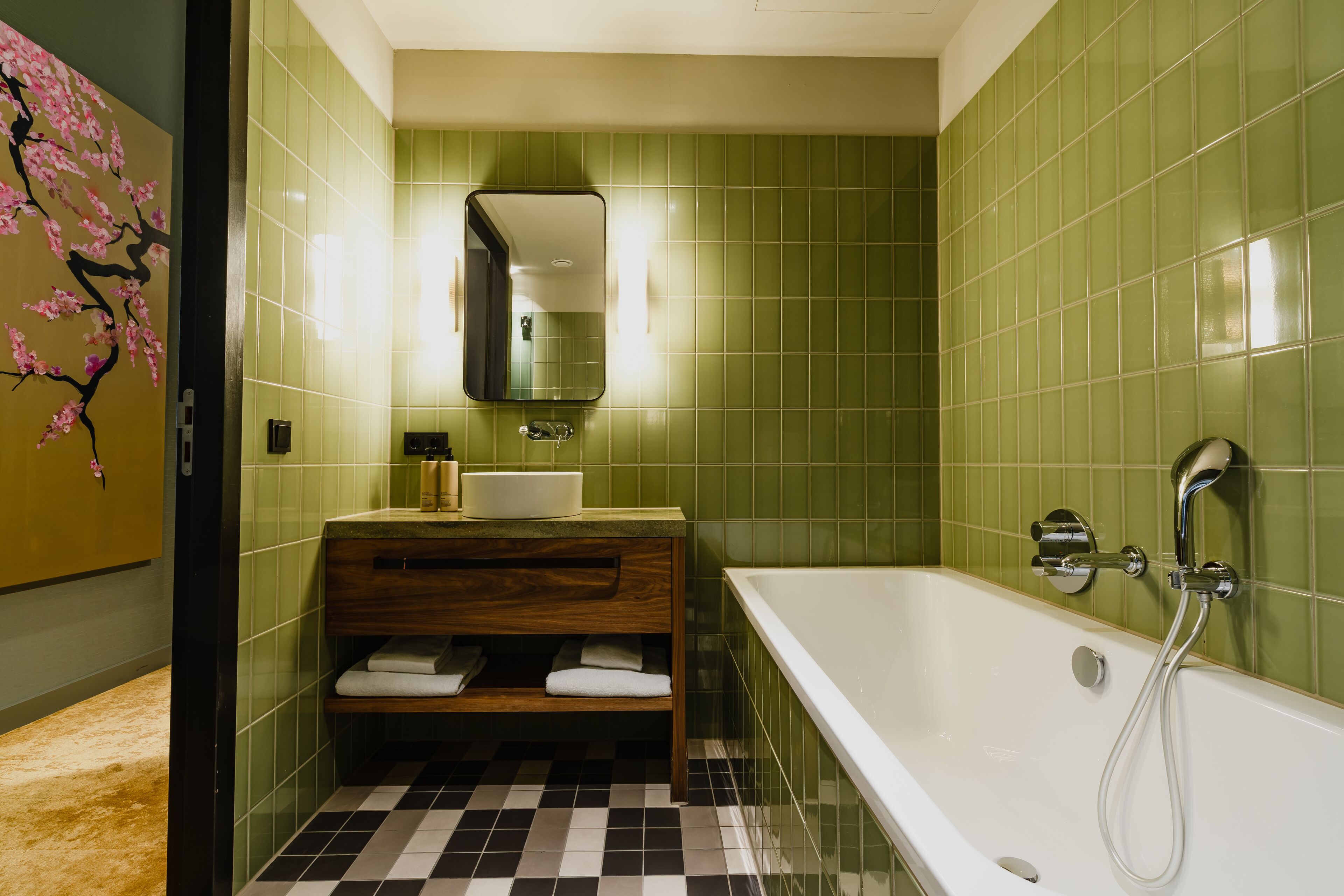 A bathroom with green tiles, a wooden vanity, a white bathtub, and a mirror above the sink.