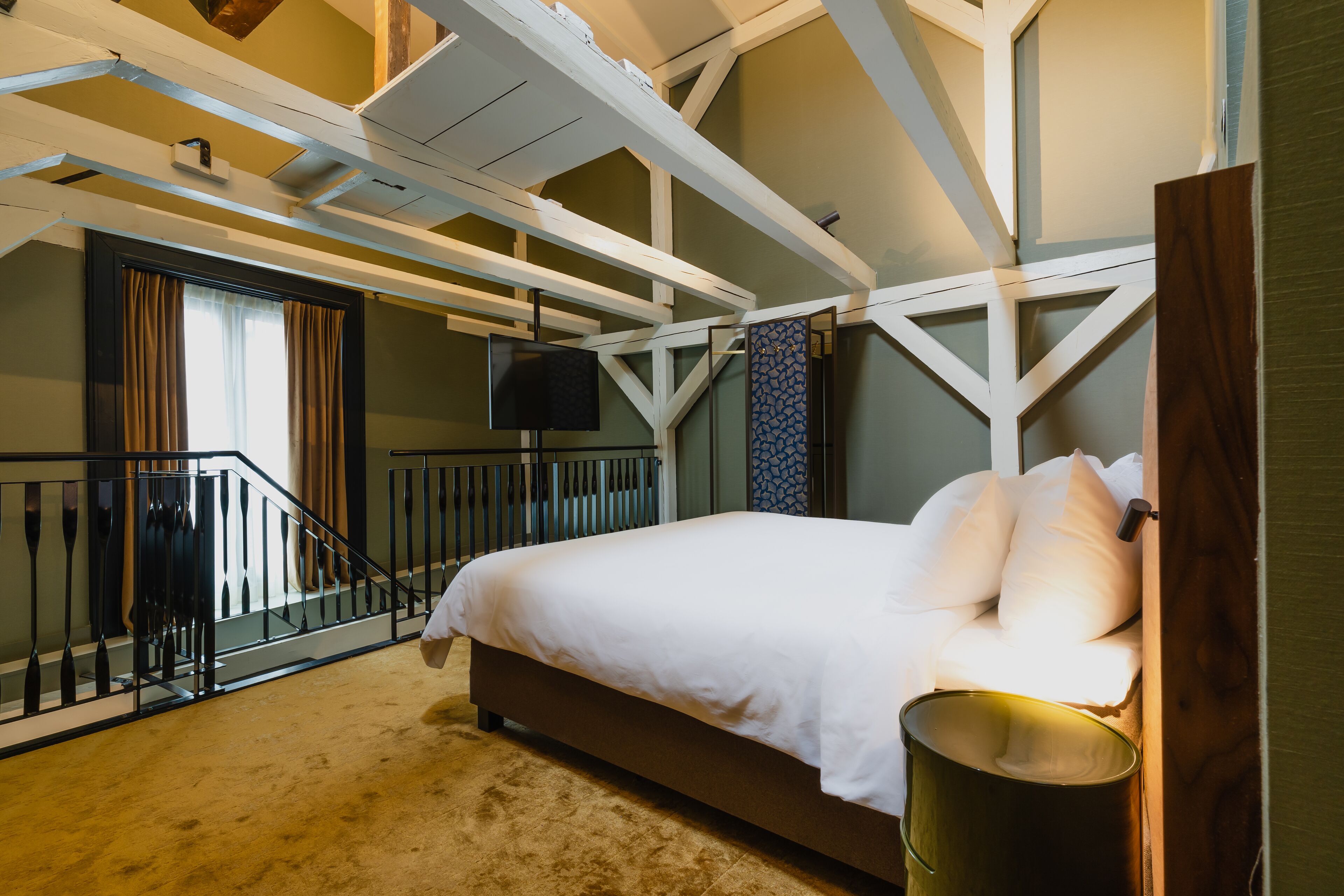 A bedroom with a bed, a wooden headboard, a window with curtains, a staircase, and a wall-mounted TV.