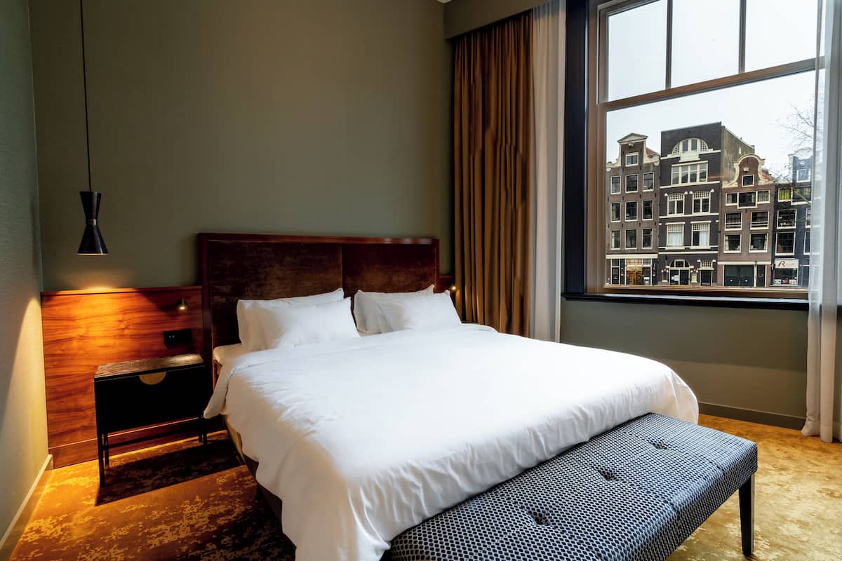 Ein Hotelzimmer mit einem großen Bett, einem Nachttisch aus Holz, einer Bank und Blick auf eine Stadtkulisse durch ein Fenster.