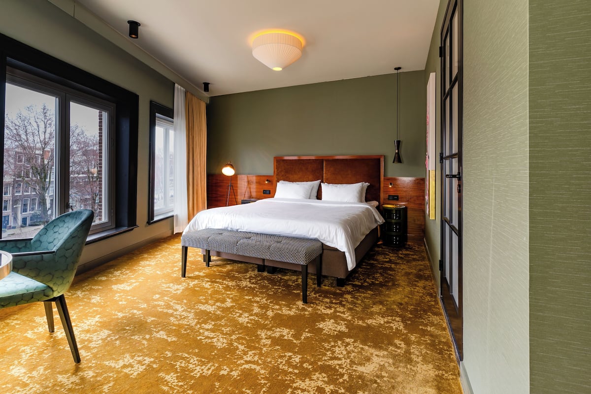 Ein Hotelzimmer mit einem großen Bett, einer Bank, einem Sessel, einem Nachttisch und Blick auf die Stadt durch große Fenster.