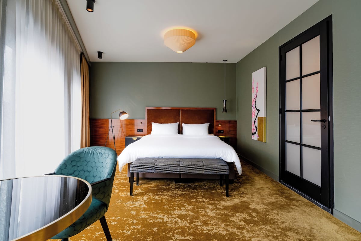 Ein Hotelzimmer mit einem großen Bett, einem Nachttisch, einem Sessel und Blick nach draußen durch ein Fenster.