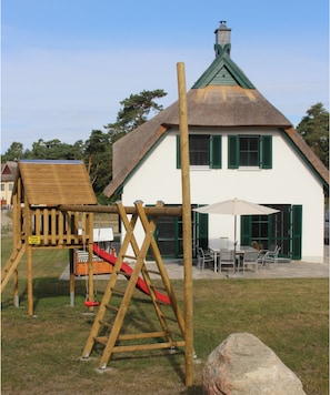 Eine Holzspielanlage mit Rutsche und Schaukeln, ein Picknicktisch und ein Gebäude mit grüner Dachbegrünung und Fensterläden.