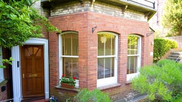 Uma casa de tijolos com porta de madeira, três janelas com persianas e um pequeno alpendre com plantas em vasos.