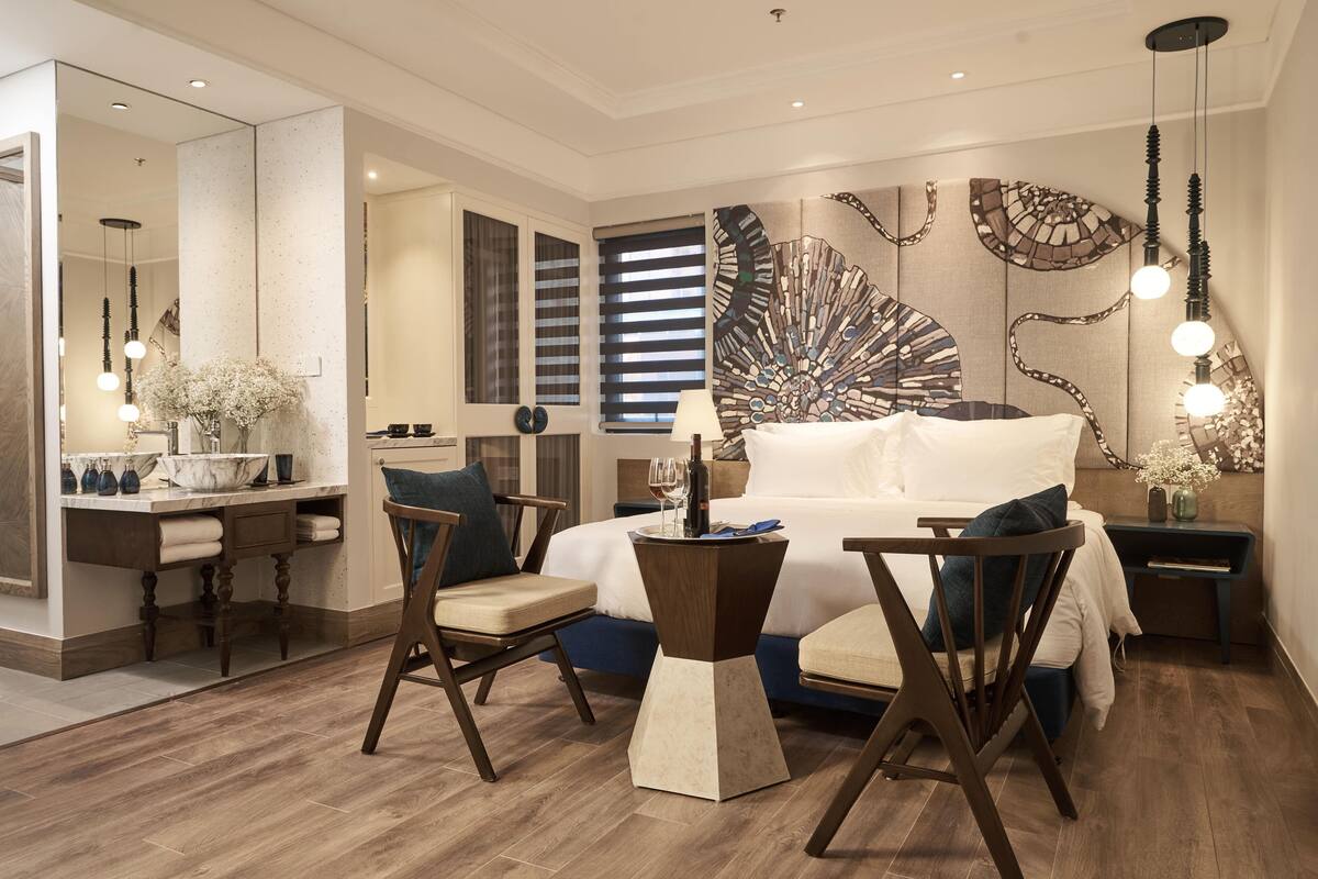 Ein modernes Hotelzimmer mit einem großen Bett, zwei Stühlen, einem Nachttisch, einem Spiegel, einem Fenster mit Jalousien und einem Wandbild.