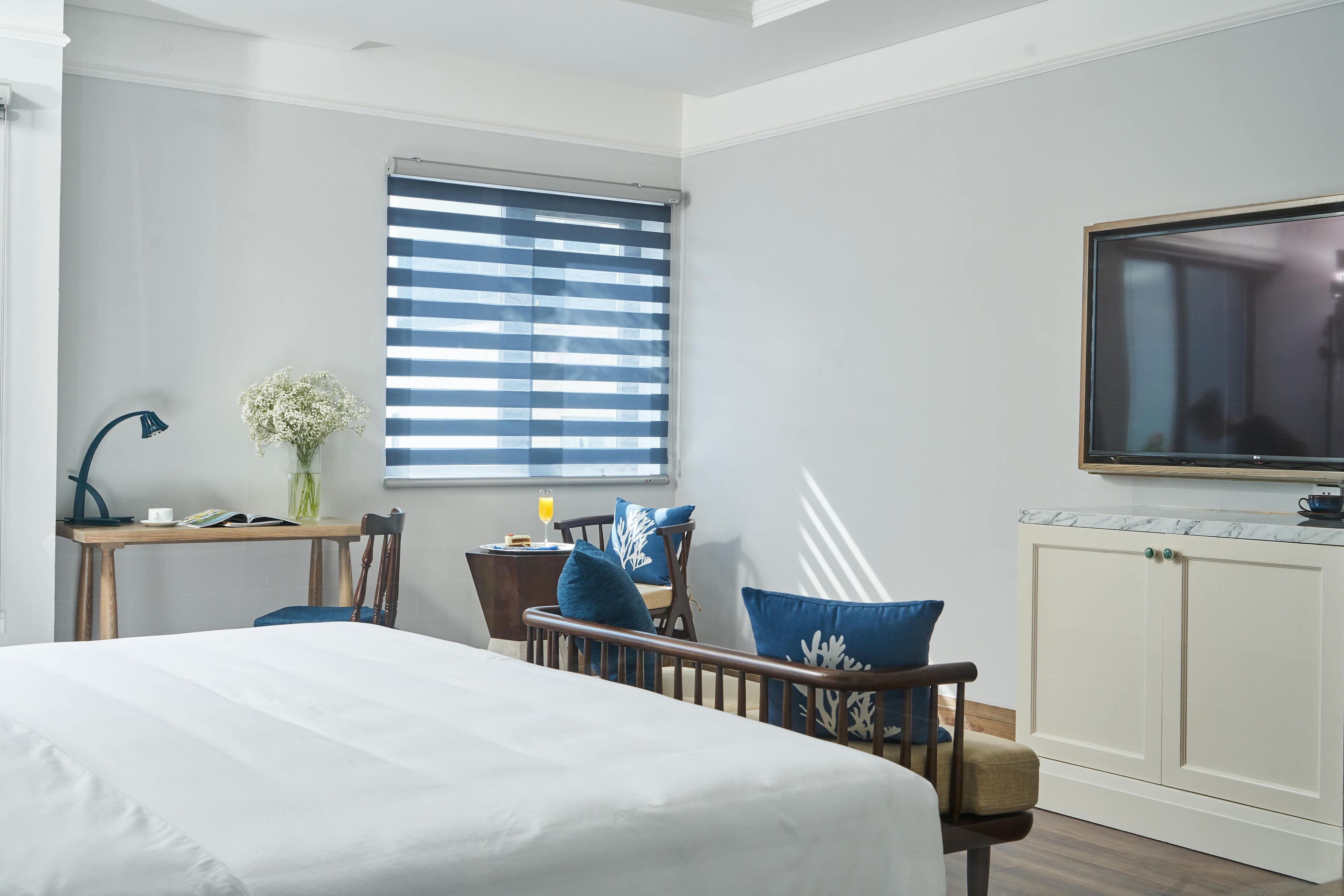 A hotel room with a bed, a desk with a lamp, a chair, a window with blinds, and a TV mounted on the wall.