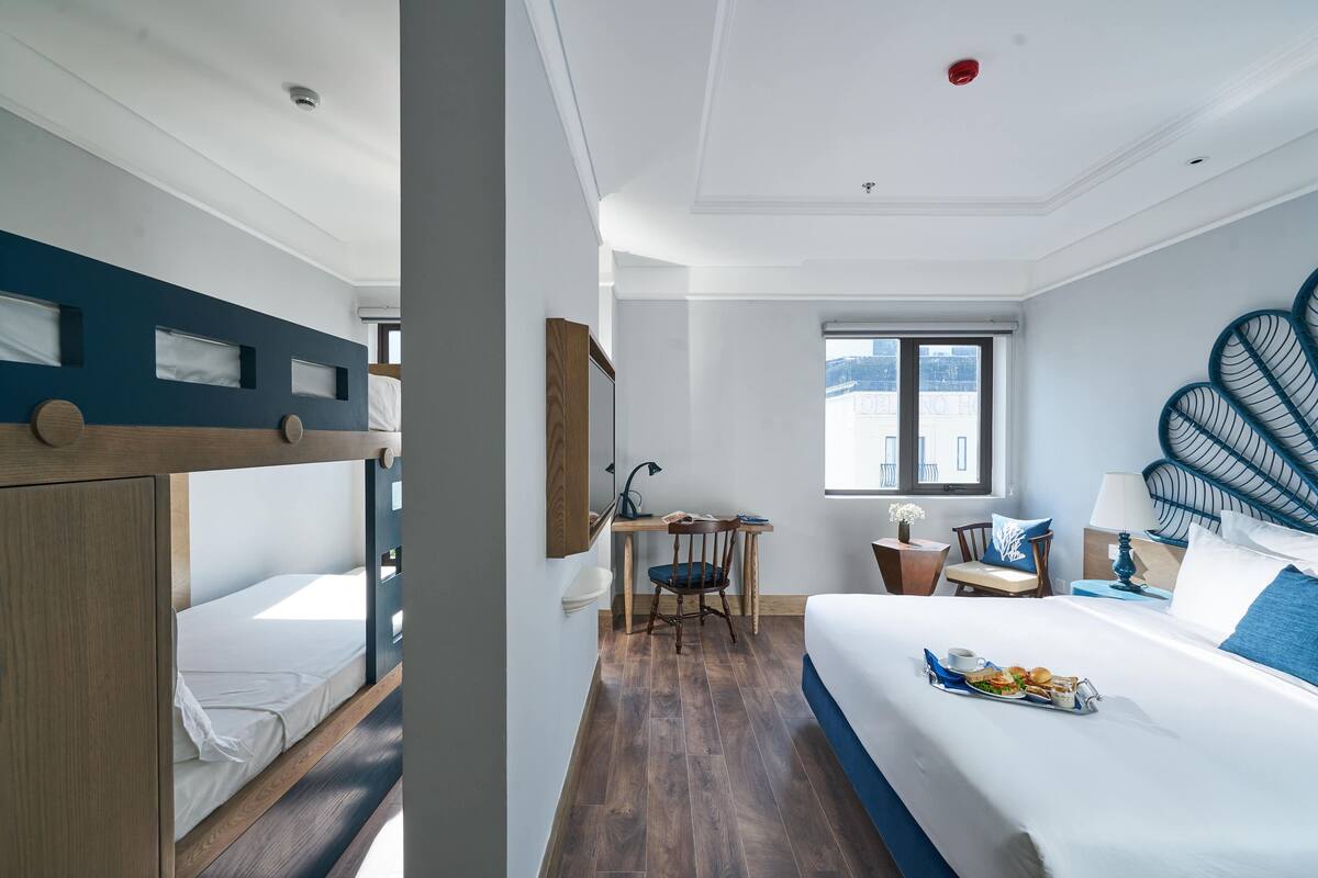 Ein Hotelzimmer mit Stockbett, Schreibtisch mit Stuhl, einem Esstisch und einer Anrichtung mit Speisen sowie einem großen Bett mit dekorativer Kopfteil.