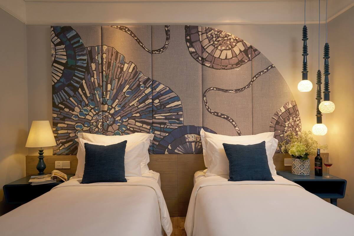 Ein Hotelzimmer mit zwei Betten, einer großen abstrakten Wandkunst, einem Nachttisch mit Lampe und einer Vase mit Blumen.