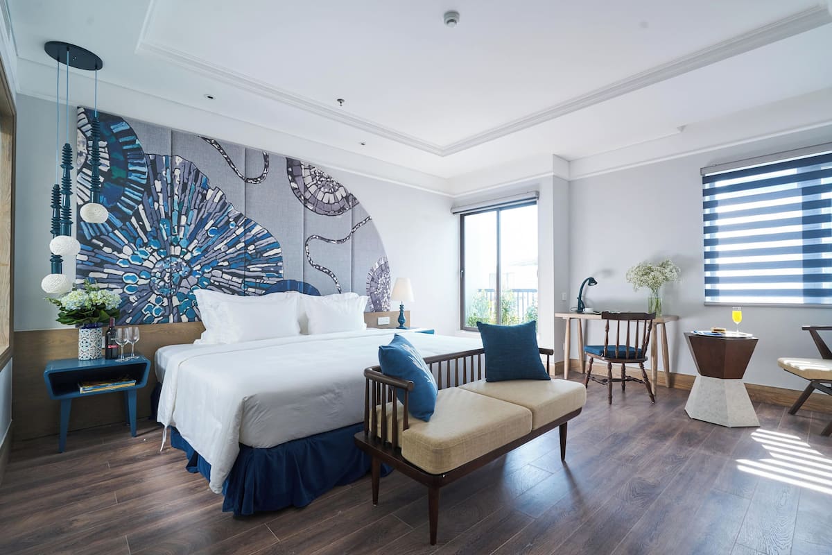 Ein modernes Hotelzimmer mit einem großen Bett, einer Couch, einem Schreibtisch und einem Sessel. Ein großes Fenster mit Jalousien, ein Deckenventilator und eine dekorative Wandgestalt sind ebenfalls vorhanden.