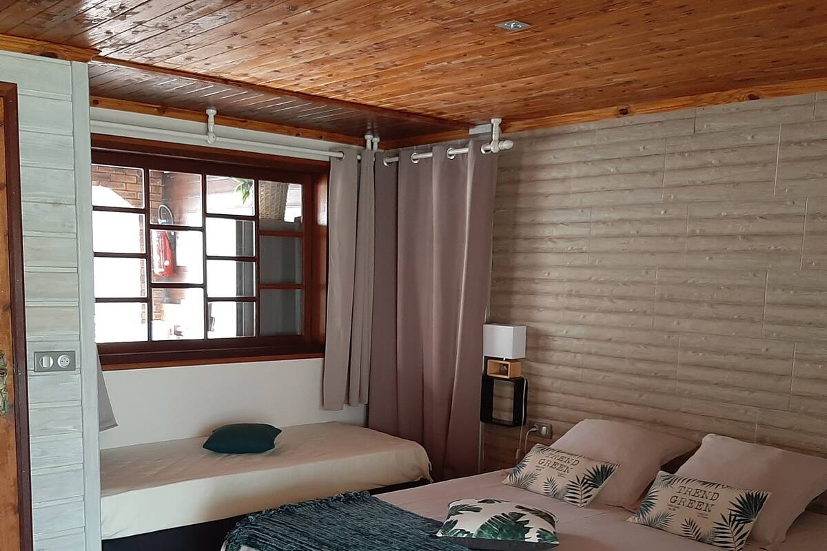 Un dormitorio con techo de madera, una ventana con cortinas, una cama con manta verde y una mesita de noche con lámpara.
